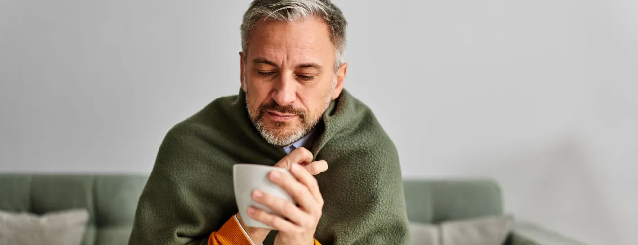 Un homme enrhumé est emmitouflé dans une couverture et a une tasse à la main.